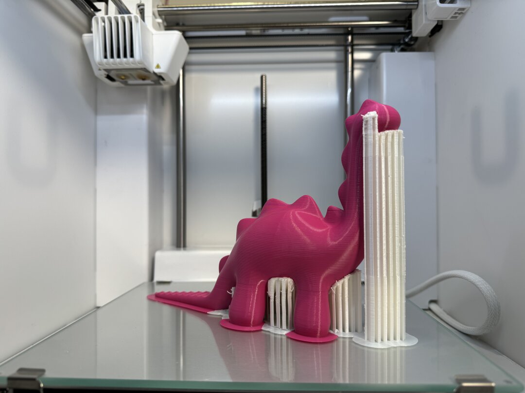 Ein Dinosaurier wird mit Stützmaterial gedruckt Auf dem Druckbett eines 3D-Druckers ist die Hälfte eines rosa Dinosauriers zu sehen. Weißes Stützmaterial umgibt ihn.