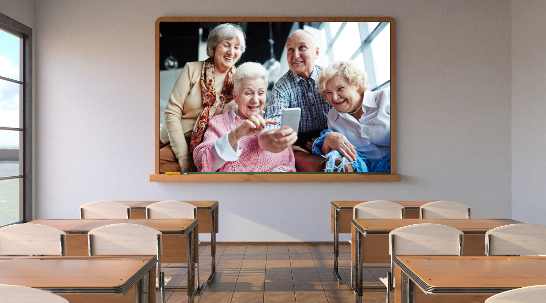 In Smartphonekursen für Senioren werden die Funktionen Schritt für Schritt erklärt In einem Klassenzimmer ohne Schüler hängt eine Tafel. Auf dieser Tafel ist ein Bild mit 3 Seniorinnen und einem Senior, die lachend in ein Smartphone schauen.